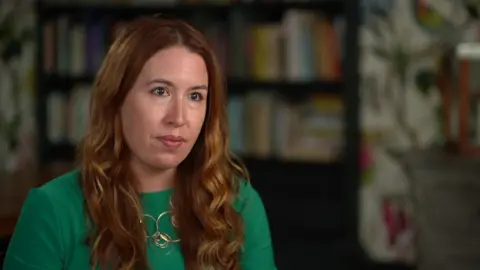 BBC Emily Hunt has long auburn hair and is wearing a green top. She is speaking to a journalist who is off camera as she sits in a sitting room that is out of focus in the background.