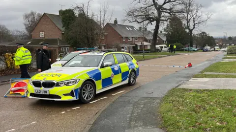Two police cars parked on a residential street. Two officers in uniform are stood beside the cars. More officers can be seen working in the background beside a line of police tape.