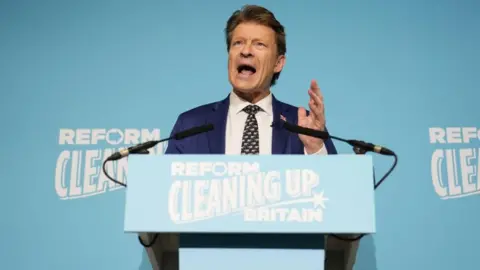 Richard Tice is stood at a lectern and is speaking to reporters, who are not pictured. The phrase "Reform Cleaning Up Britain" is written on the lectern and on the background wall. Tice is wearing a blue jacket, white shirt and patterned tie.