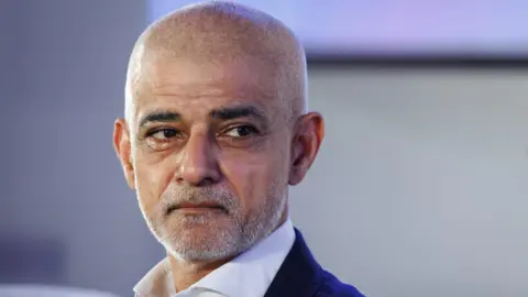 Getty Images Close-up portrait of Sadiq Khan. He has has a shaved and a neat beard, wearing a white open-collared shirt and a dark suit jacket against a blurred background.