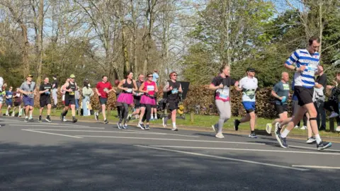 Crowds of people including some women in brightly coloured dresses warn over their fitness attire running along a road with spectators on the grass and pavement beside it.