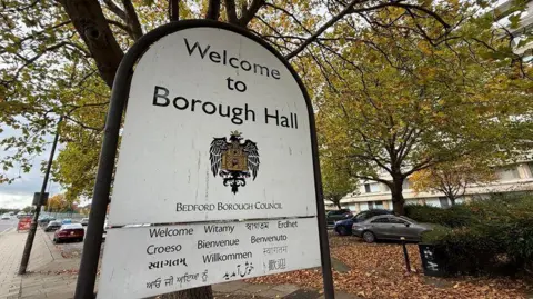 Steve Hubbard/BBC A large Bedford Borough Council entrance sign with "Welcome to Borough Hall", a crest and "welcome" in several languages, all in black lettering. It is hung from a black frame and there are trees behind it, with much leaf-fall on the ground. There are parked cars in a car park beyond the sign.