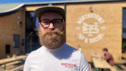 Mousumi Bakshi/BBC Sam Adams is standing off centre to the left, and is looking at the camera. He has a long ginger beard and moustache, a brown hat, black glasses, and a grey T-shirt. 