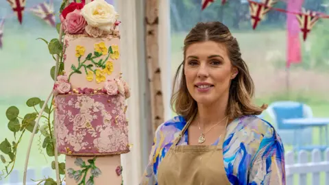 Georgie Grasso stood in the Bake Off tent. She is wearing a beige apron and underneath it has a blue, pink and white marbled top. She is looking at the camera with her brown and blonde hair half tied up and half down. There is a cake to the left of her, which is a three tiered cake, in different shades of pink with flowers adorned on it. 