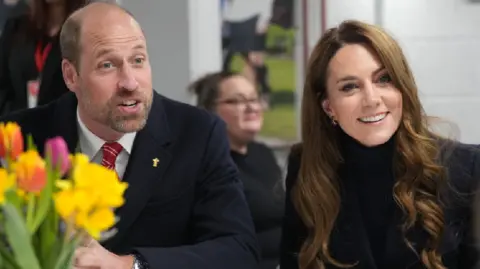 PA Media The Prince and Princess of Wales in the Sir Tasker Watkins Suite. Both aren't looking at the camera. William is on the left and is talking to someone, whilst Kate is on the right and smiling. In the left corner there are a bouquet of flowers, including daffodils. There is a woman blurred in the background. 