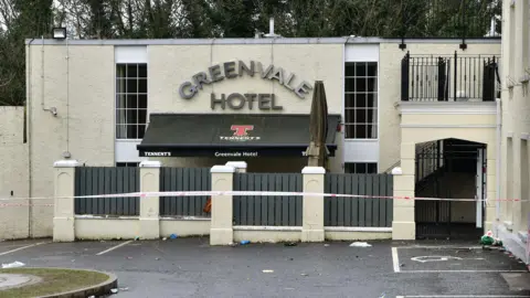 Pacemaker The exterior of the Greenvale Hotel in Cookstown in the aftermath of the fatal crush in March 2019. Only part of the building is visible and it consists of a flat-roofed, two-storey, cream rendered structure, with a gated entrance below a gated balcony area. A large grey fence with cream pillars shields the front of the building from a small car park which is strewn with a few pieces of litter. A black, branded awning protrudes from the building behind the grey fence. A red and white police tape cordons off the scene. 