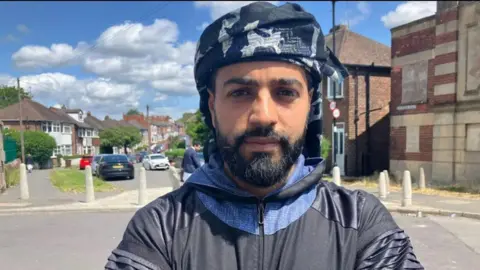 BBC/Phil Bodmer Saleh Alsirkal stood in the street with a serious expression. He has a black beard and black hair and is wearing dark clothing.