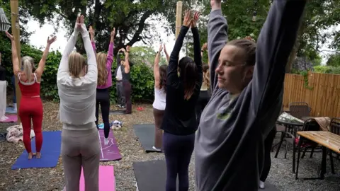 Connor Bennett/BBC A group standing on yoga mats with their arms stretching into the air, one in the foreground has a grey jumper on and her eyes closed