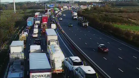 A motorway with vehicles stuck in long queues on one side. There are cars and trucks in the queues.