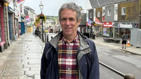 A man with grey hair wearing a black, white, and yellow plaid shirt and dark blue rain jacket with Market Jew Street in the background.