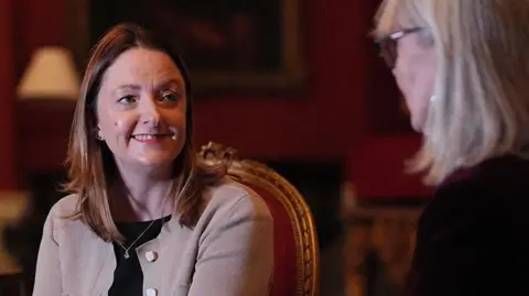 Kristina Penny with long brown hair wearing a beige smart cardigan with a black top. She is in Number 10 sitting on an ornate gold and red velvet chair talking to her aunt who has long blond hair wearing a black outfit to the right.