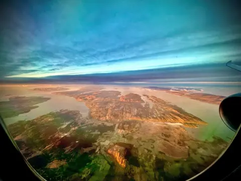 Anthony Parker Aerial view through an airplane window showing rugged land, water, and islands below, with soft light across the landscape and a wide, cloudy sky above.