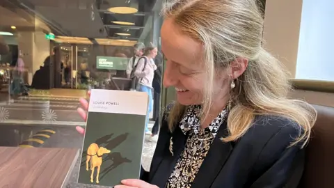 Anna-Marie Fitzgerald Louise Powell holding a copy of Underdogs on the left and looking down at it. The cover is green with a yellow drawing of a greyhound with the shadow of a man and boy behind the dog. There is a strip of white at the top with the writing Louise Powell and then Underdogs beneath it. Powell has her hair tied back at the top and is wearing a black and white patterned shirt underneath a black blazer. She is sitting in front of a glass wall with what looks like an office behind it.
