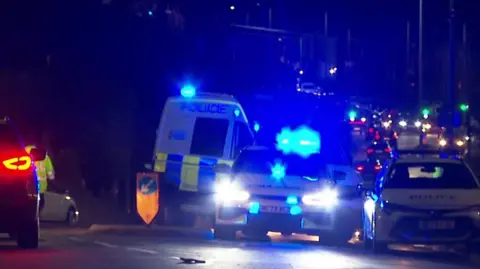 Emergency service vehicles including a police van and cars on Bolton Road in Ashton-in -Makerfield in front of a line of traffic. It is dark.