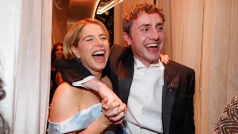Jessie Buckley, in a blue strapless dress, and Paul Mescal, in a tuxedo, laugh with their arms around each other