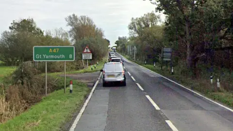A stock image of the A47 Acle Straight showing cars backing up. The road is one lane in either directions. A road sign showing Great Yarmouth is on the side of the road, which is surrounded by fields and shrubbery.