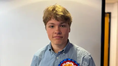 Gabriel Raimondo who aged 18 became a deputy in Jersey. He has blonde hair and is wearing an unironed blue shirt with a red, blue, white and yellow rosette on his left chest.