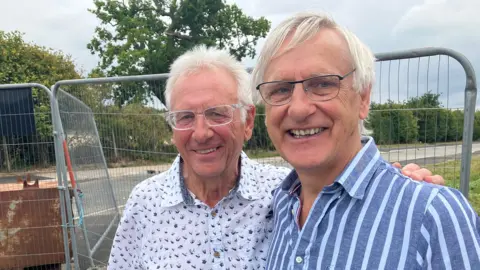 BBC Two former male doctors wearing glasses smiling and standing in front of metal fencing 