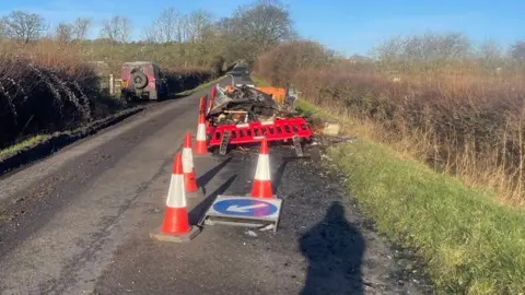 Philip Spicer A pile of mostly indistinguishable rubbish in a narrow rural road, which has been coned off. A keep left barrier has collapsed alongside the cones, there is also a red barrier leaning against the rubbish. There is a grass verge to the right alongside a hedge, with fields beyond.