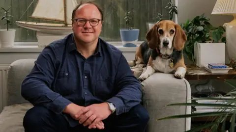 Russell Hunt Russell is sitting on a grey armchair beside a brown, white and black beagle in a cosy, plant‑filled living room. 