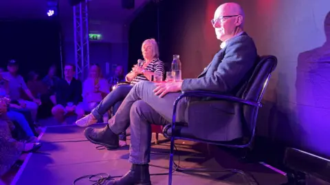 PA Media John Swinney and Susan Morrison, both dressed casually, on stage. the lighting is very purple.