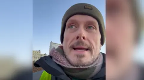 A man in his fifties wearing a scarf and hat. He is standing on the beach. 