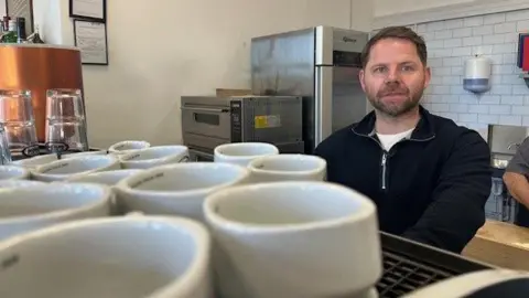 Chris Allen stands behind a coffee machine with mugs piled on top of it. He has short brown hair and a beard.