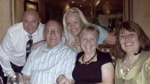 FAMILY HANDOUT Mr and Mrs Eccleston are sitting on chairs next to a woman with brown hair. Behind them is Ms Munn, their daughter, and a man. They are all smiling at the camera.