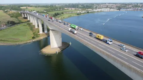 John Fairhall/BBC An aerial view of the Orwell Bridge showing the southern bank of the estuary. Traffic is queuing on the eastbound carriageway, where two lanes are open as vehicles approach the lane closure. Traffic is flowing on the westbound carriageway, where both lanes are also open. A marina and south Ipswich are in the top right of the image. A road passes under the bridge on the left of the picture.
