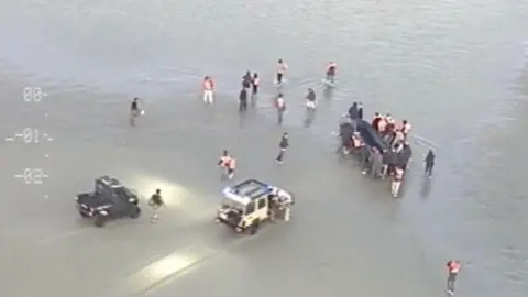 Two cars in the shallow water with people surrounding what appears to be a dingy boat with people inside the vessel