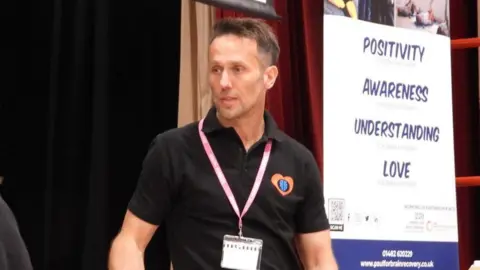A man in a black polo shirt with a orange heart logo with blue icons inside is wearing a pink lanyard and looking to the side. He has short brown hair and next to him is a poster which says "positivity, awareness, understanding" and "love" on it.