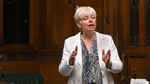 Lia Nici when she wa an MP speaking in the House of Commons she has short blond hair and is wearing a multi-coloured shirt and a whire jacket