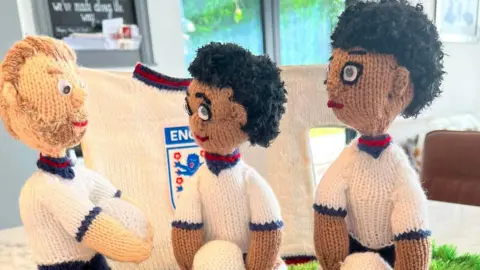 Jan Platt Three woollen figures depicting England football players. Each of them are holding a white ball. There is a knitted white England shirt behind them.