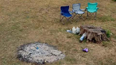 Three camp chairs, one green, one blue and one grey are next to a tree stump and the remains of a campfire. There are bottles, cans and wrappers littered on the dry looking grass.