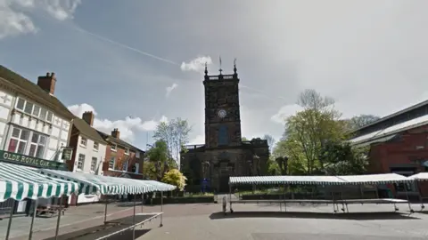 Google A town centre with a church in the centre of the image and market stalls in the foreground. There are pubs and shops surrounding an open public square.