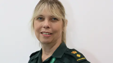 South Western Ambulance Service A blonde woman in a green uniform that says 'paramedic' on the shoulder