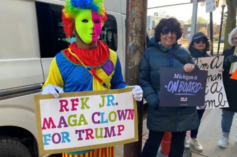 BBC / Mike Wendling A man dressed in a clown suit and holding a sign saying "RFK Jr - MAGA clown for Trump"