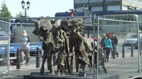 Thomas Nugent/Geograph Sculpture, apparently in bronze, showing a family carrying large suitcases. The work is surrounded by metal fencing. Pedestrians and cars are visible behind the structure.