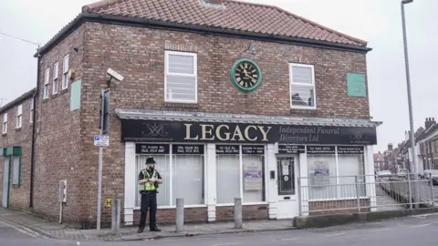 PA Media A police officer stands outside a branch of Legacy funeral directors. The building's entrance is straight on to the street and sits between two roads, forming T junctions on both sides of it.