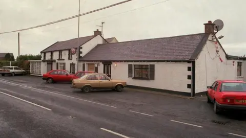 Pacemaker A cottage style building with white washed walls and cars from the 1990s parked outside. 