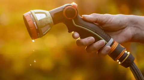 Close-up of a hand holding a garden hose spray nozzle, with water droplets falling from the nozzle against a blurred golden background.