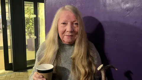 Holly Nichols/BBC A woman with long, wavy hair wearing a grey knitted sweater. She is holding a disposable coffee cup in one hand and a walking stick with a curved handle in the other. The background features a deep purple wall and part of a glass entrance door with visible reflections and some indoor plants behind the glass. 