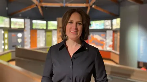 Helen Glencross is smiling. She is standing in a museum and display boards can be seen in the background.
She has brown hair and is wearing a black blouse with a small pin broach on her lapel.