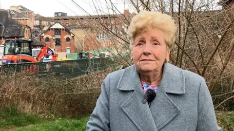 Resident Tina Conway-Jameson is an older woman with short blonde hair, and   wearing a grey coat while stood on some grass with the bridge in the background.