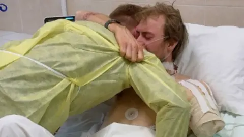 Base UA AD and one of their fellow volunteers in a hospital room in Ukraine. AD's shoulder is closed after dissection of his hand. He is hugging fellow volunteers tightly.