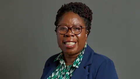 Paulette Hamilton pictured against a grey background. She is wearing a green pattered shirt underneath a blue blazer and dark-framed glasses. Her dreadlocks are pinned in a high-side bun.