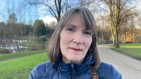 Nicky looking into the camera, she's wearing a blue puffer coat. Behind her is Shrewsbury Quarry, a park with open green space, trees, and a path stretching off into the distance. To her right is the River Severn. It's a sunny, wintery day and you can tell the light is beginning to fade. 