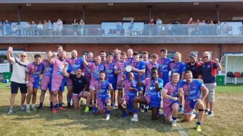 Henry Miller/BBC The Anglian Vipers squad pose for a team photo pitchside with the East Rugby League trophy as fans, friends and family look on from a balcony behind them