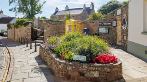 States of Alderney A stone flower bed blooming with greenery and colorful roses sitting in the middle of a stone area near a cobbled street and yellow phone box.
