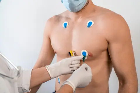 A shirtless man has electrodes attached to his chest by a doctor for a test detecting early symptoms of cardiovascular disease